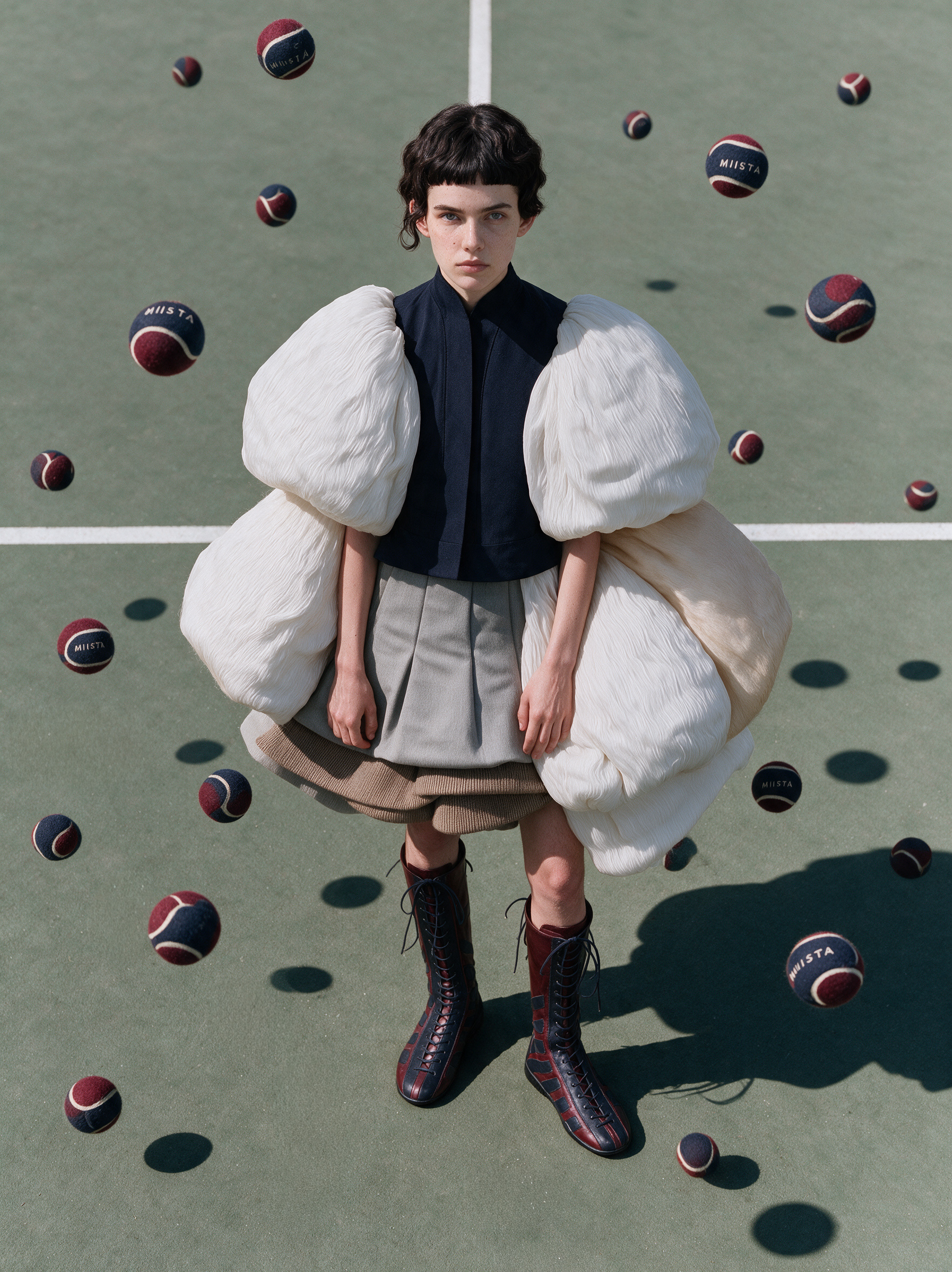 Model in sculptural white sleeves on a tennis court among floating MIISTA tennis balls